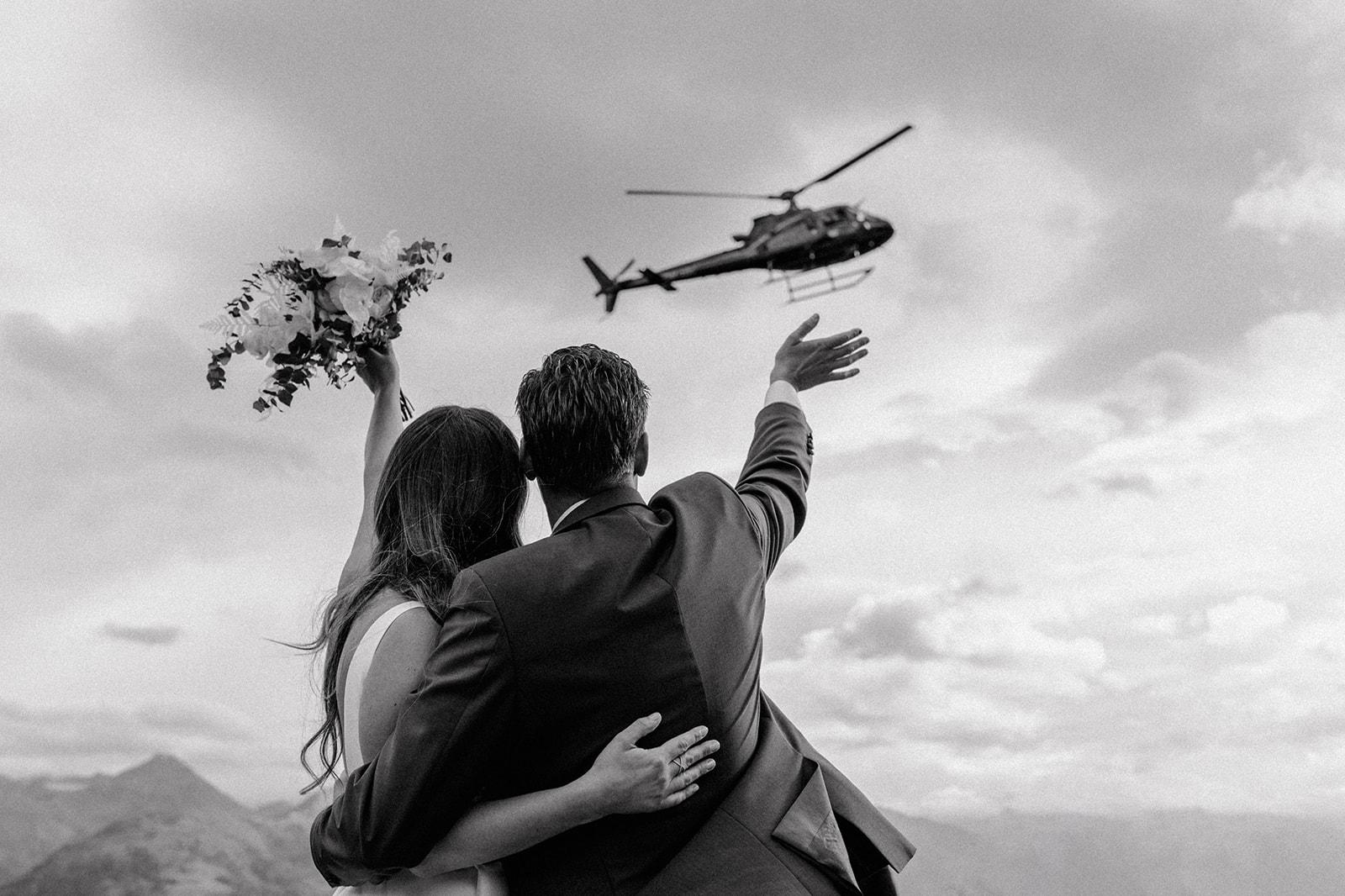 Brautpaar steht Arm in Arm da und winkt dem wegfliegenden Helikopter hinterher